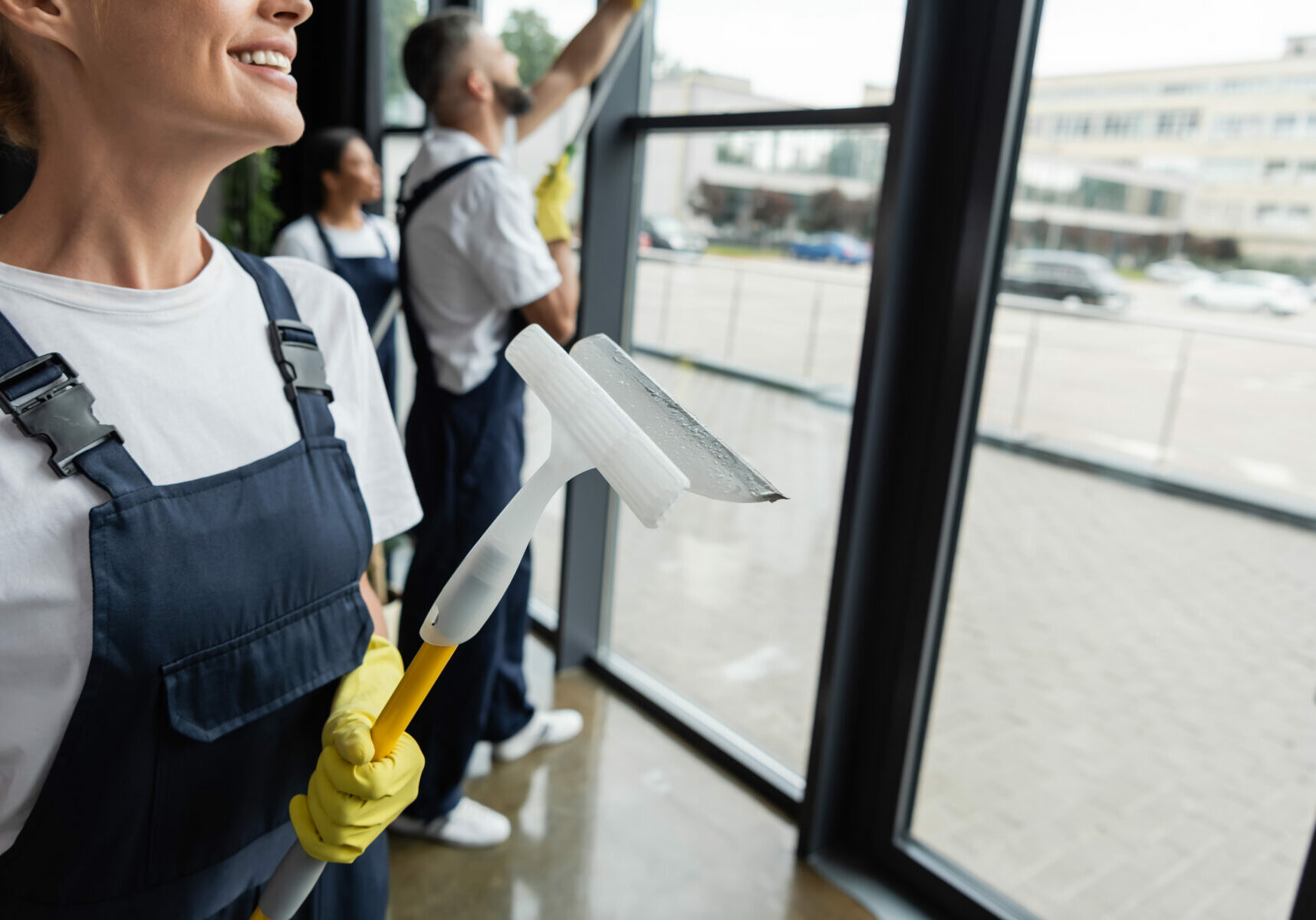 smiling woman in overalls holding window squeegee near blurred multiethnic colleagues