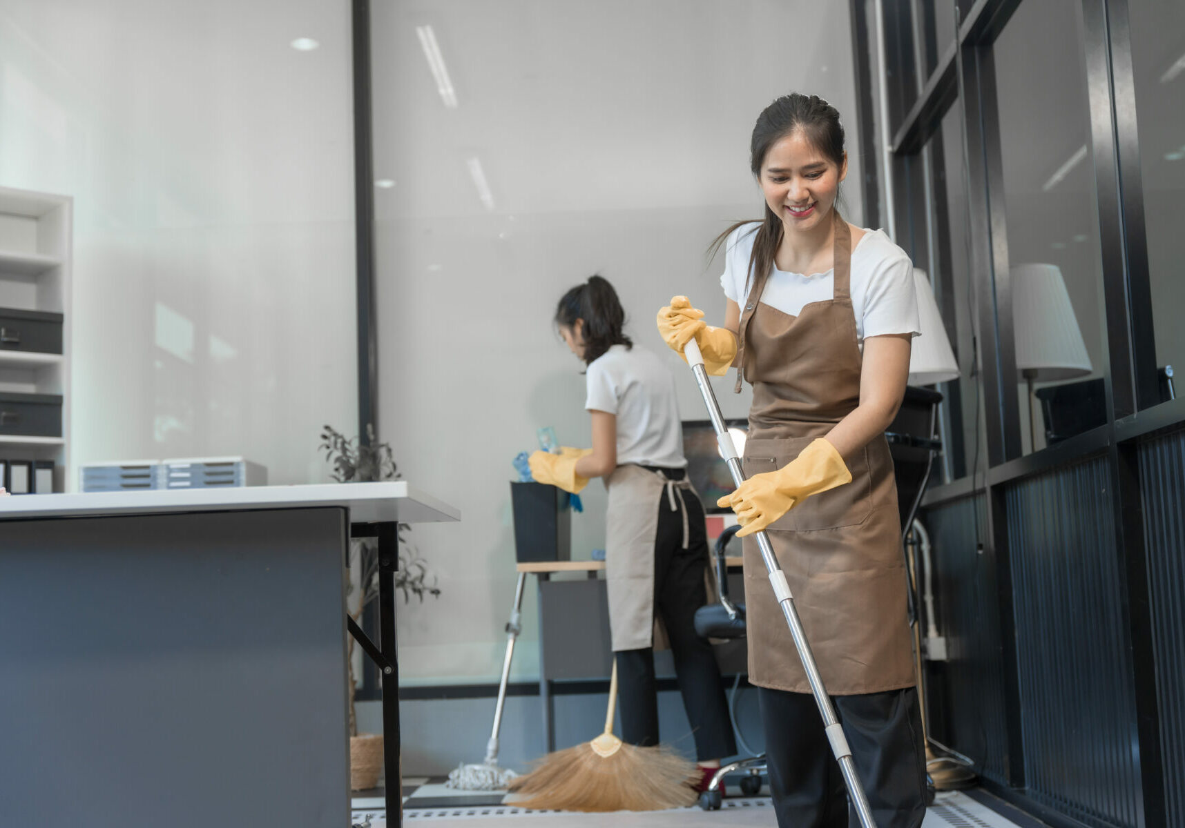 Two Asian housekeepers wear overalls and work together efficiently to clean the living room, study