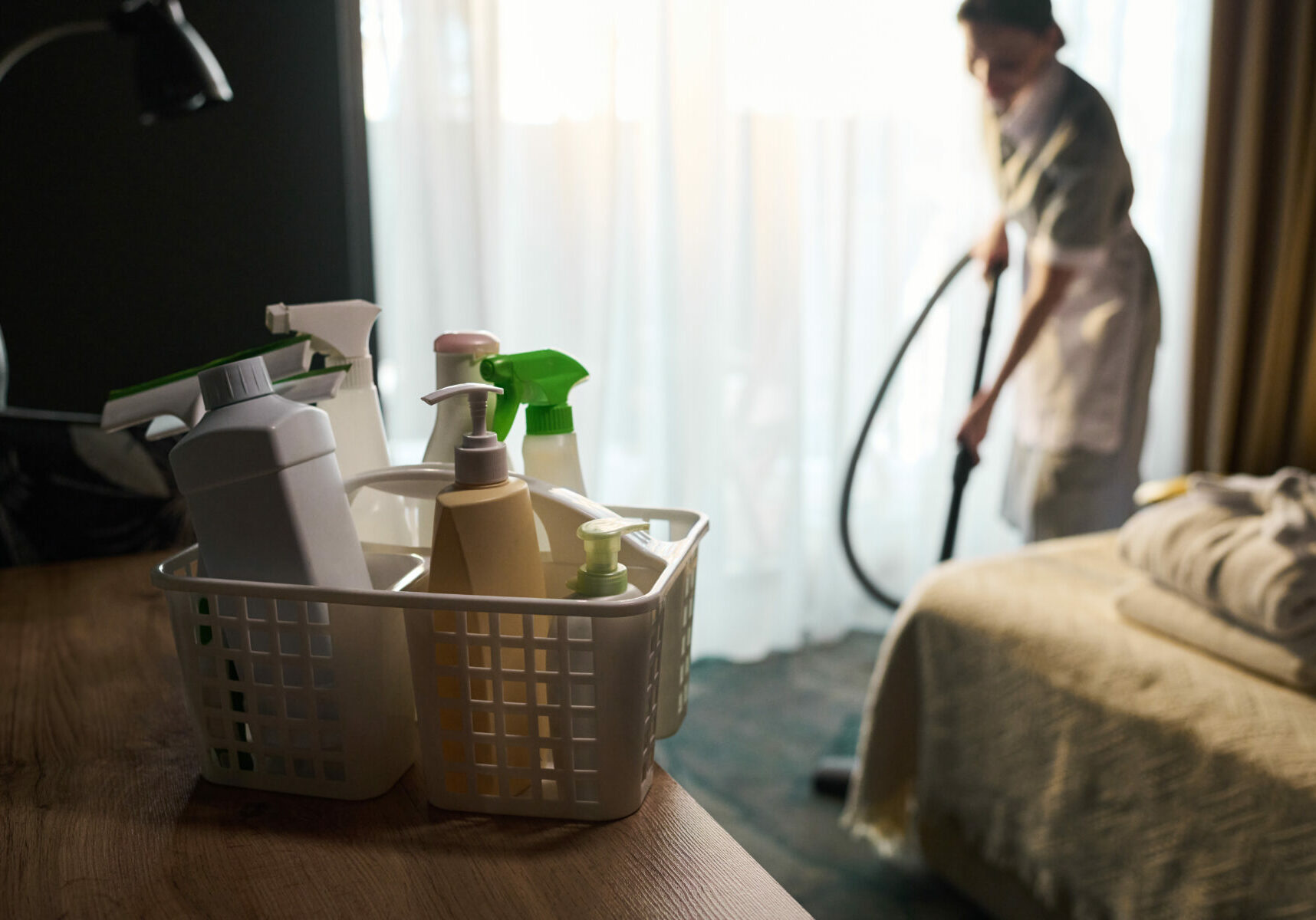 Person vacuuming room while various cleaning supplies sit on table in foreground. Bright light from window illuminates the scene