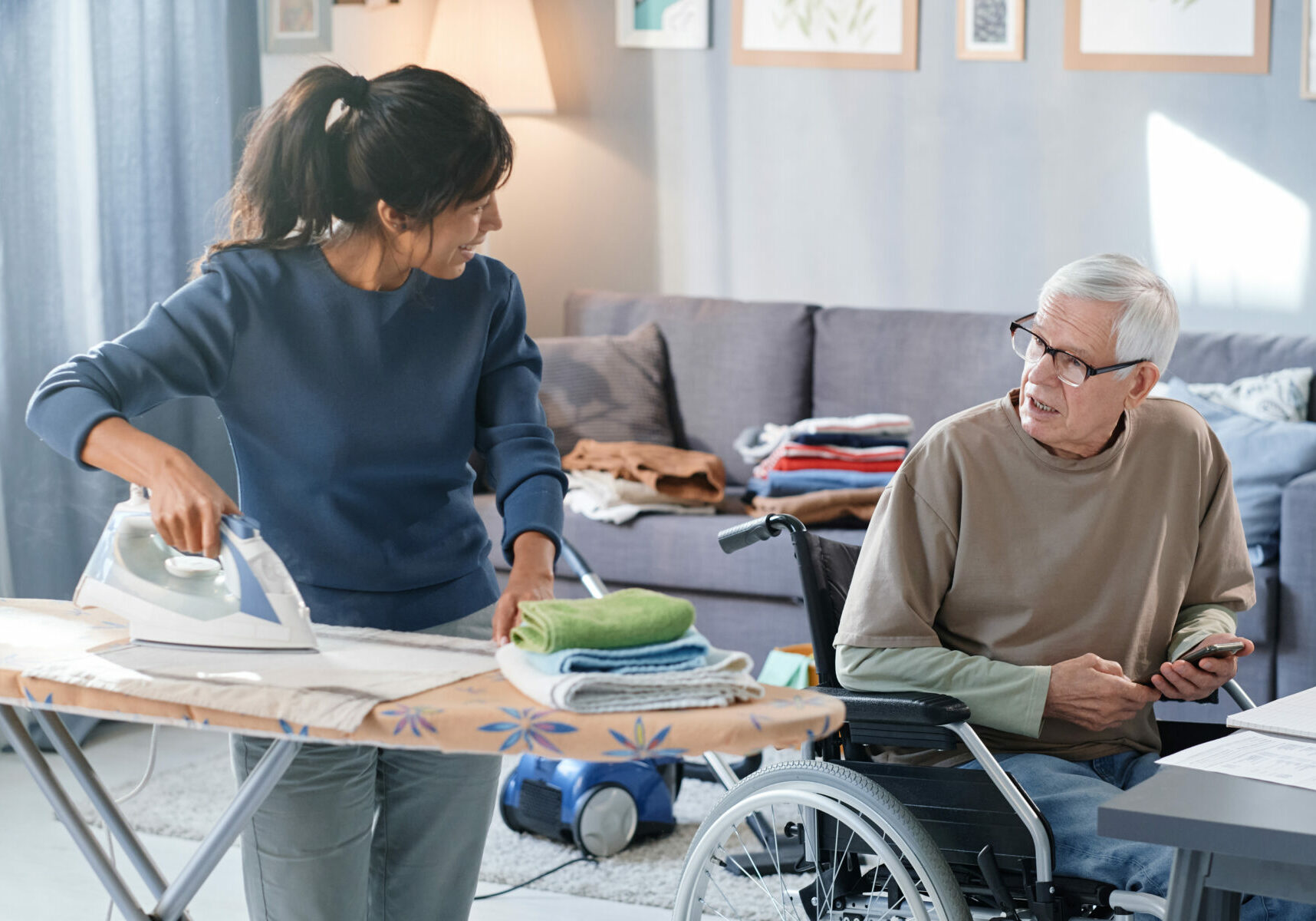 Volunteer ironing clothes and talking to senior man who using wheelchair, she helping him with chores in the room